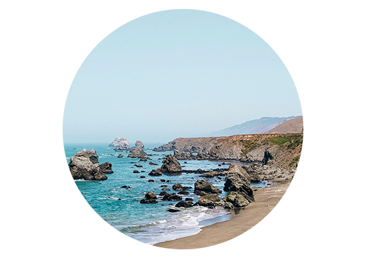 A rocky coast with shimmering ocean waves, the surf meeting the sand, with foggy mountains in the horizon.